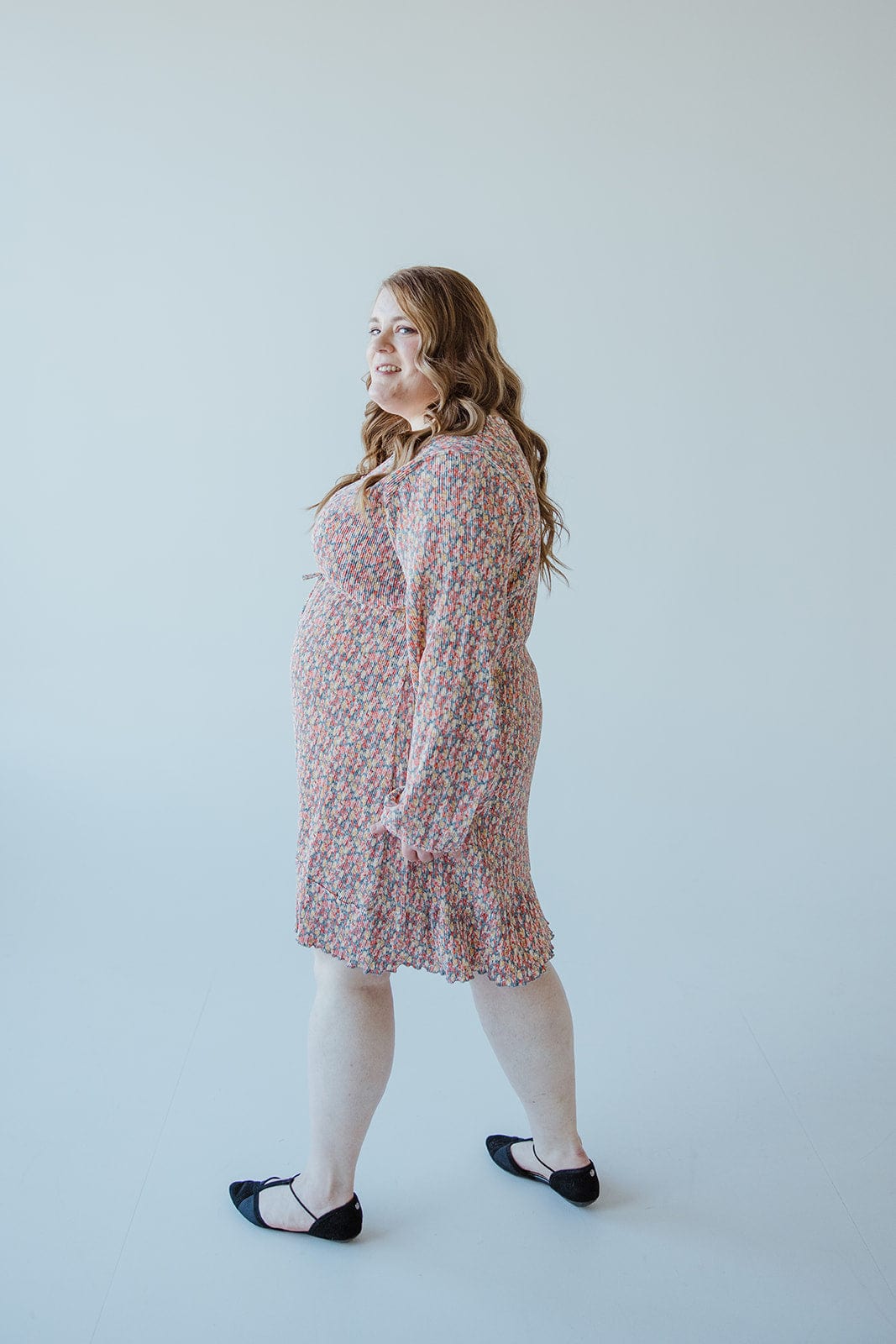 A person with long hair stands sideways, smiling, wearing a BOHO FLORAL DRESS WITH RUFFLE HEM IN FESTIVE BLOOM by Love Marlow and black flats against a plain background.