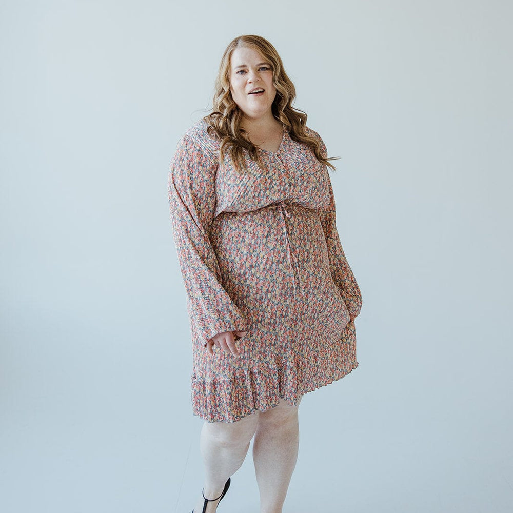 A woman with long hair stands against a plain white background, exuding boho vibes in a BOHO FLORAL DRESS WITH RUFFLE HEM IN FESTIVE BLOOM by Love Marlow and black heels.