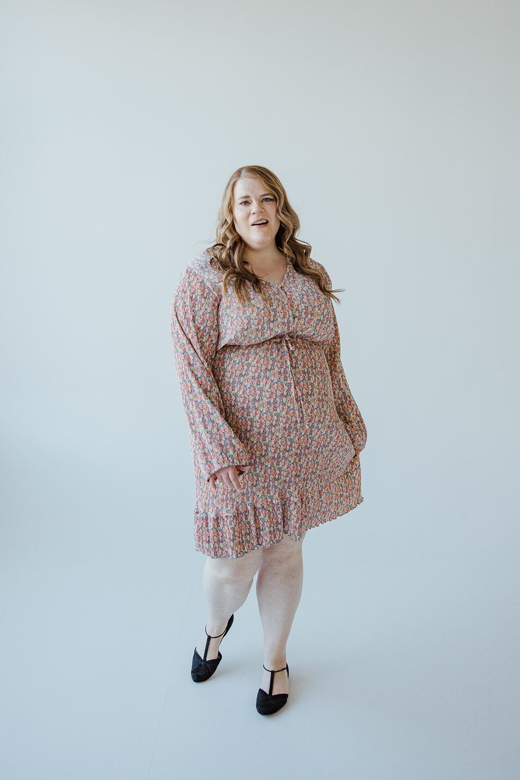 A woman with long hair stands against a plain white background, exuding boho vibes in a BOHO FLORAL DRESS WITH RUFFLE HEM IN FESTIVE BLOOM by Love Marlow and black heels.