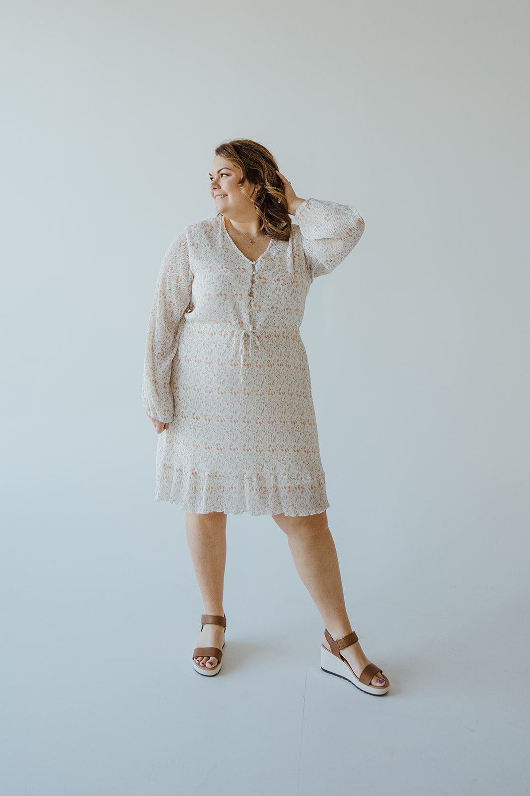 A person stands against a light background wearing the Love Marlow BOHO FLORAL DRESS WITH RUFFLE HEM IN VINTAGE OFF WHITE with long sleeves and sandals, exuding boho vibes, with one hand touching their hair.