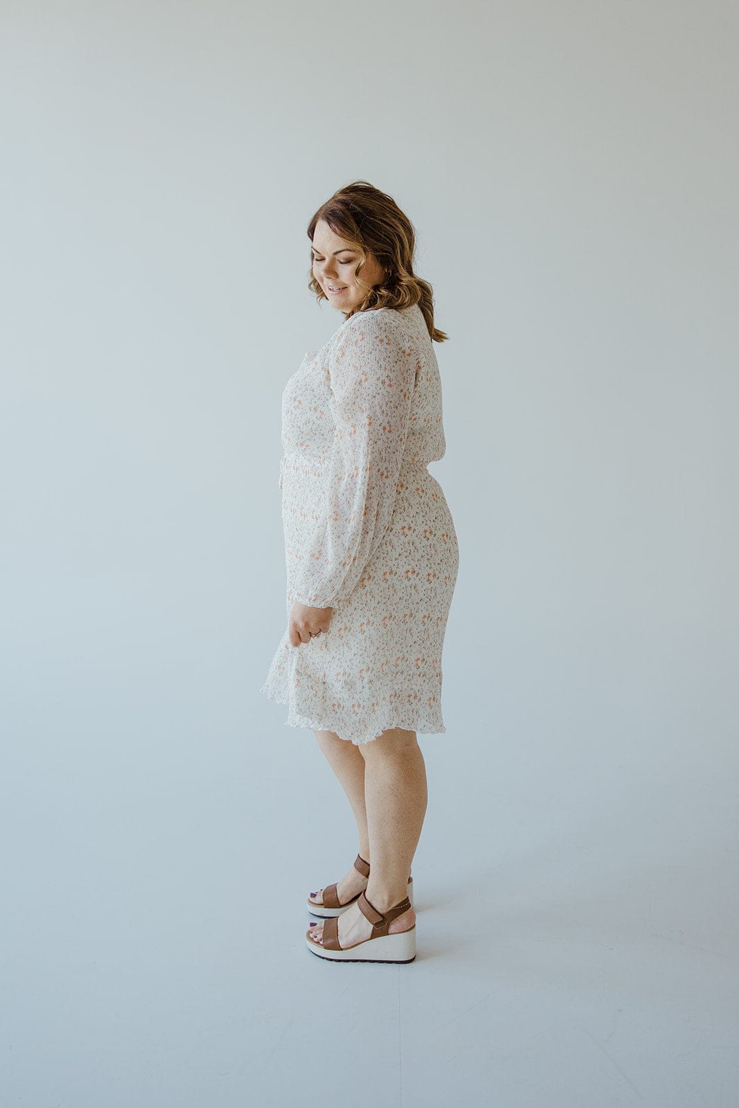 A person stands sideways wearing the Love Marlow BOHO FLORAL DRESS WITH RUFFLE HEM IN VINTAGE OFF WHITE and sandals in a plain, light-colored room.