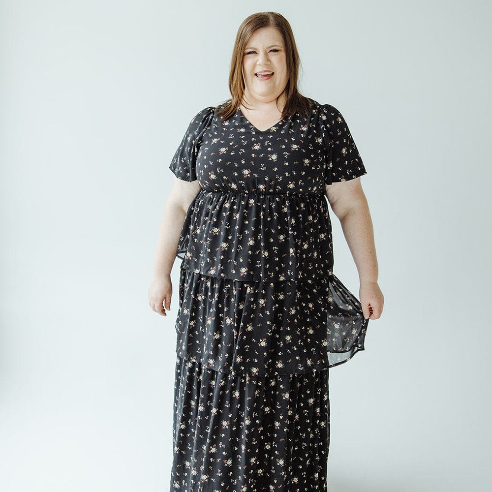 A person wearing the BOHO LAYERED RUFFLE DRESS by Mikarose stands against a plain light-colored background, smiling slightly with a hint of Bohemian style.