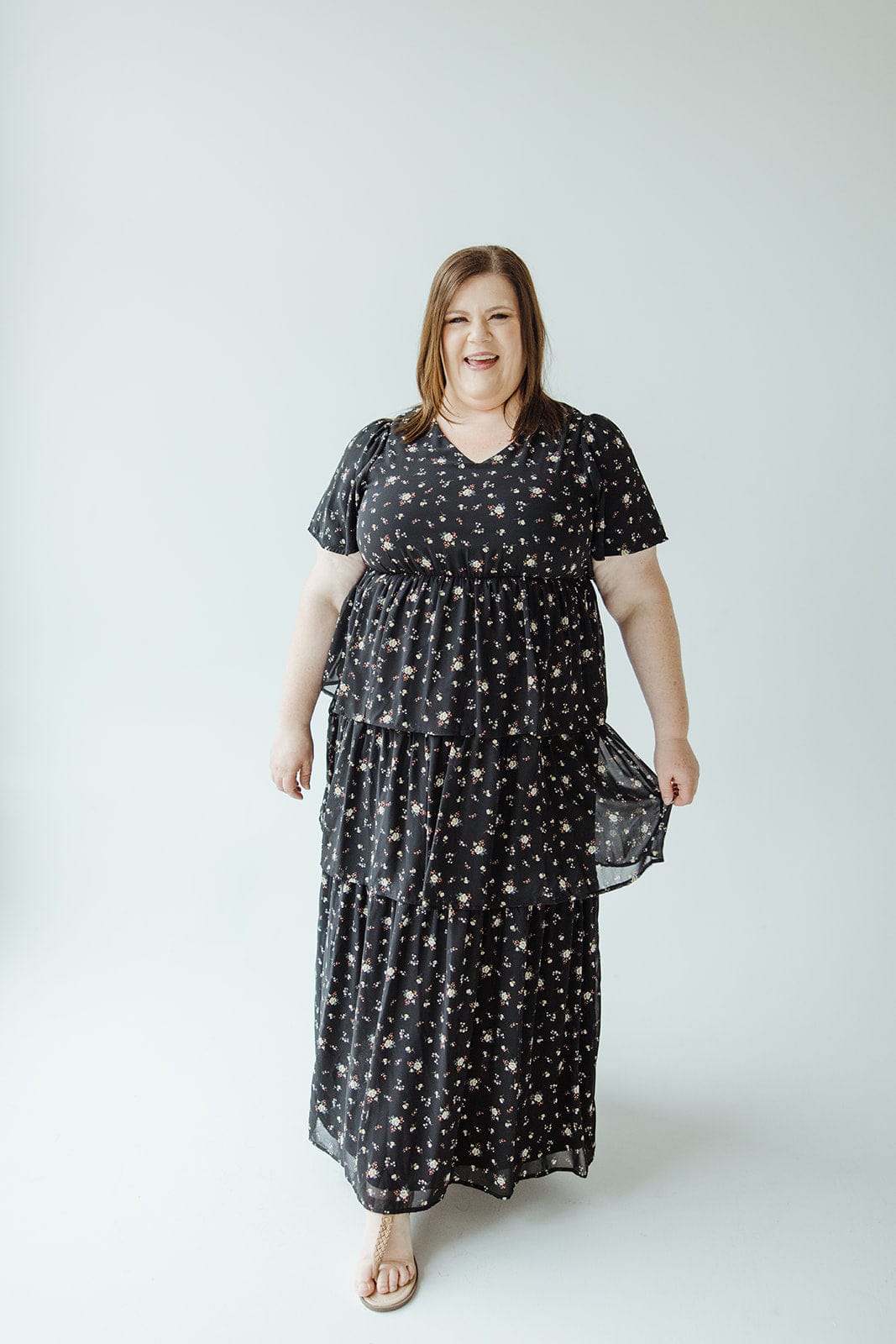 A person wearing the BOHO LAYERED RUFFLE DRESS by Mikarose stands against a plain light-colored background, smiling slightly with a hint of Bohemian style.