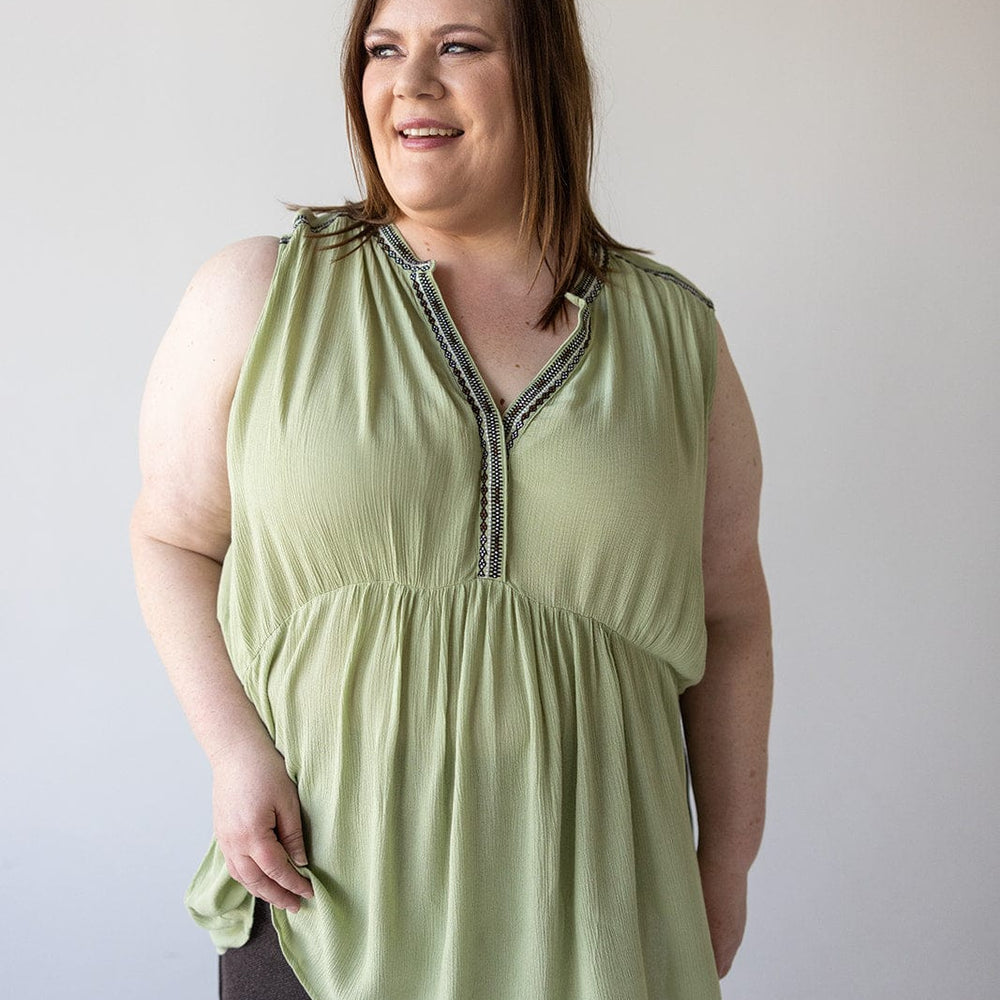 A woman stands against a plain background, wearing a light green BOHO TANK WITH EMBROIDED DETAIL by Love Marlow made of lightweight fabric and dark pants, with a slight smile on her face.