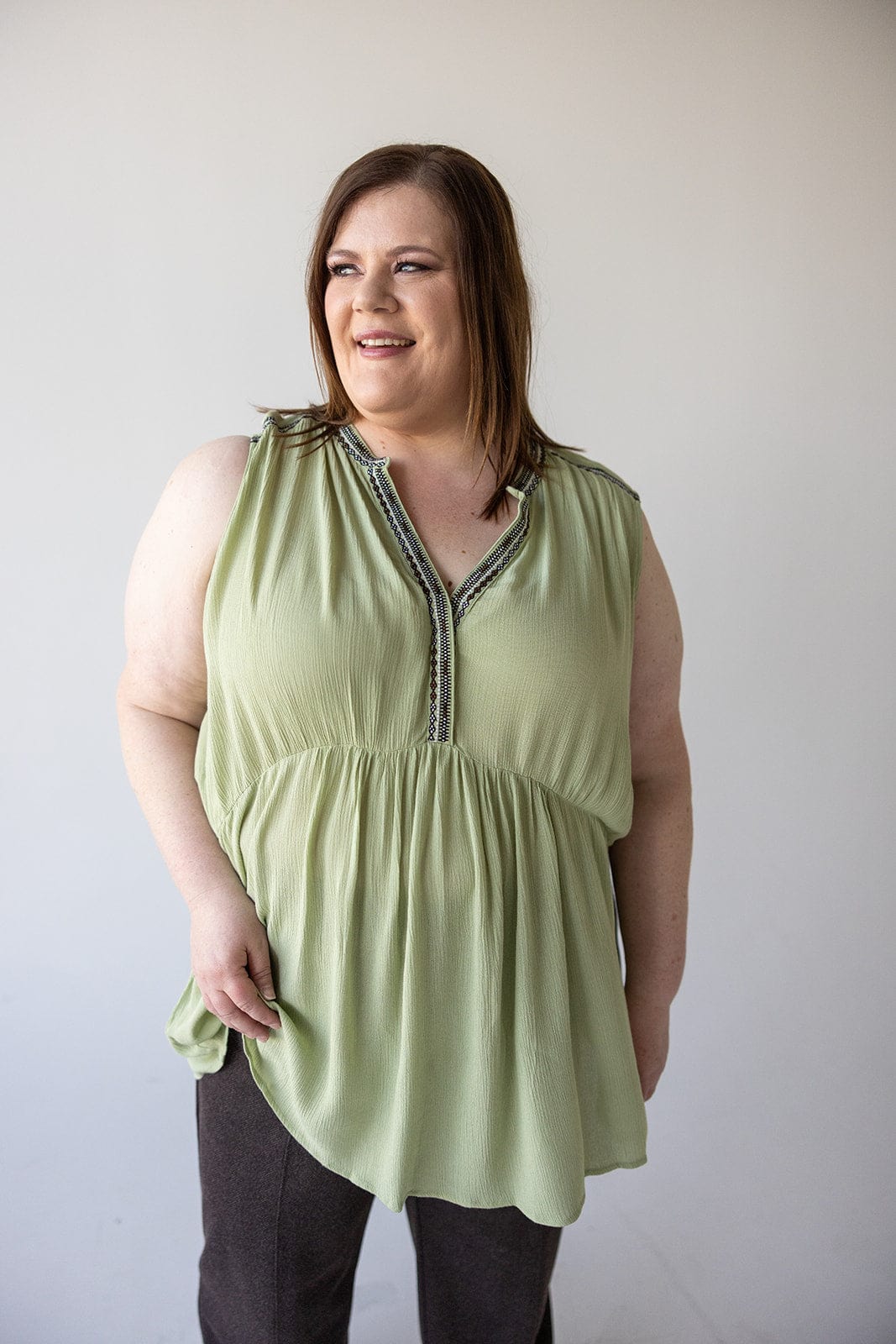 A woman stands against a plain background, wearing a light green BOHO TANK WITH EMBROIDED DETAIL by Love Marlow made of lightweight fabric and dark pants, with a slight smile on her face.