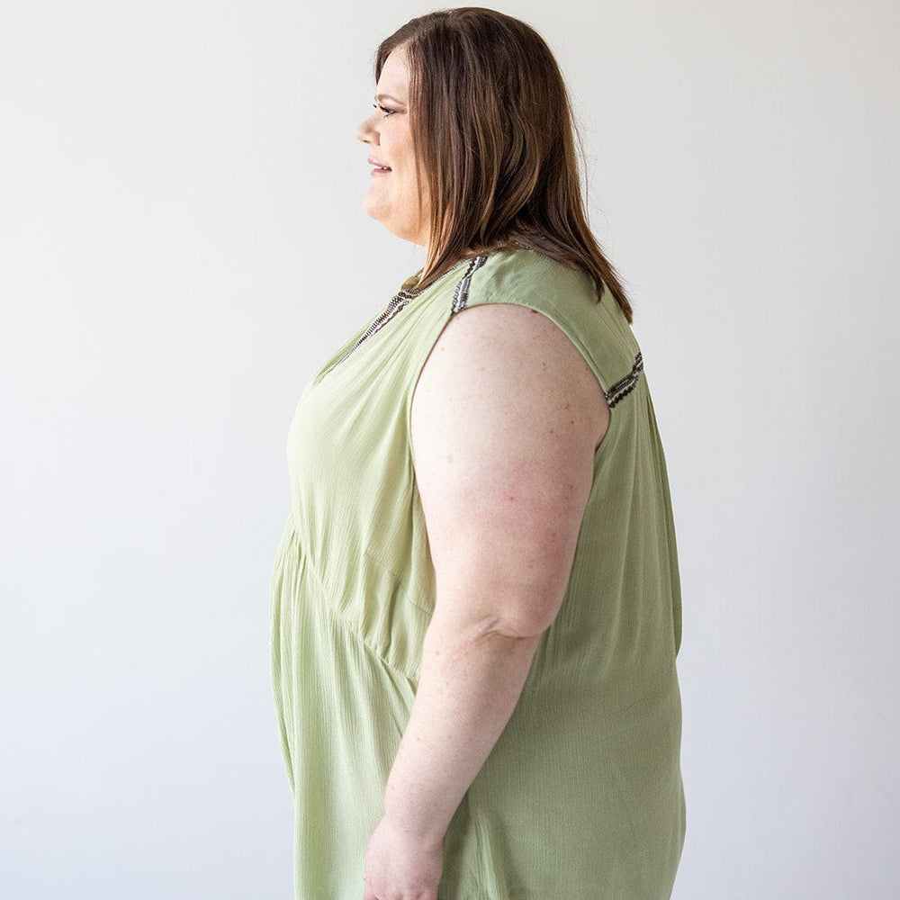 A woman with shoulder-length brown hair is standing in profile, smiling, and wearing a light green BOHO TANK WITH EMBROIDERED DETAIL from Love Marlow and dark pants against a plain white background.