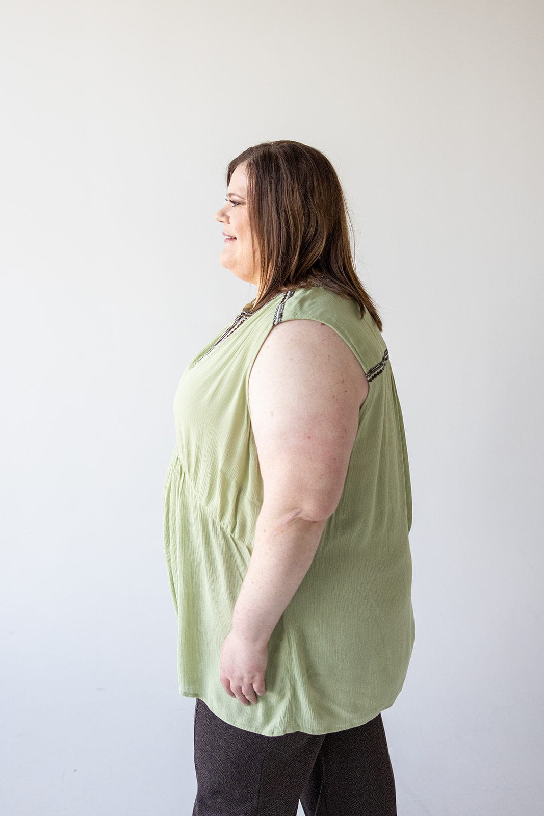 A woman with shoulder-length brown hair is standing in profile, smiling, and wearing a light green BOHO TANK WITH EMBROIDERED DETAIL from Love Marlow and dark pants against a plain white background.