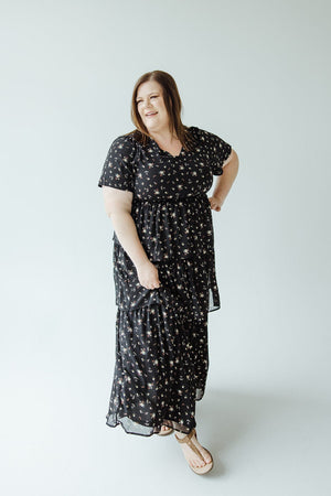 A person wearing a black Bohemian-style floral dress from Mikarose, known as the BOHO LAYERED RUFFLE DRESS, along with sandals stands against a plain white background, smiling.