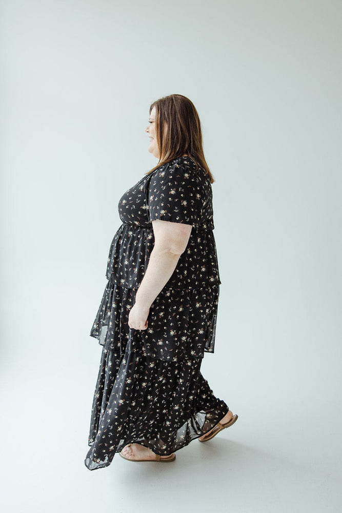 A woman in a Mikarose BOHO LAYERED RUFFLE DRESS, featuring a black floral Bohemian style, walks sideways against a plain background.