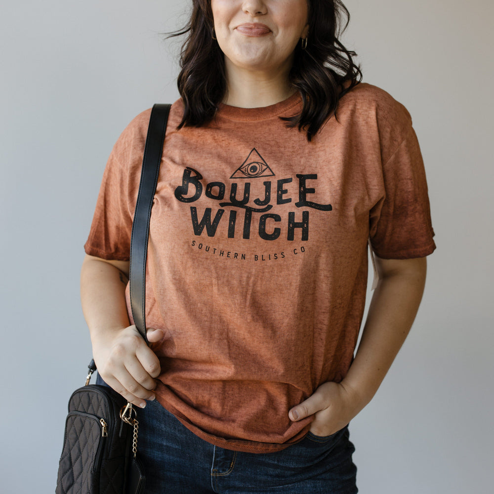 Standing against a plain background, a person dressed in Love Marlow's "BOUJEE WITCH" graphic tee—a rust-colored shirt featuring eye-catching Halloween graphics—holds a black quilted purse.