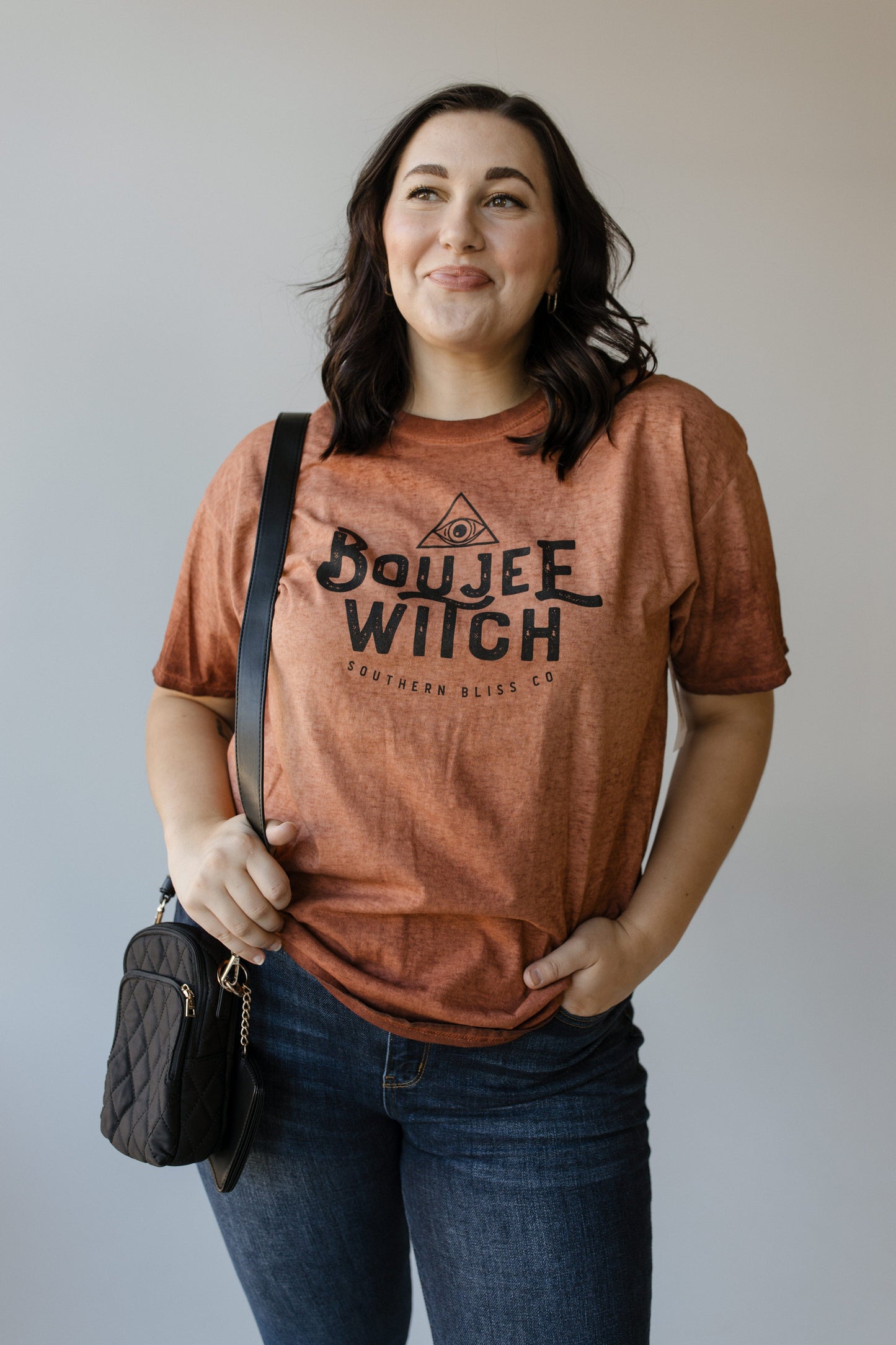 Standing against a plain background, a person dressed in Love Marlow's "BOUJEE WITCH" graphic tee—a rust-colored shirt featuring eye-catching Halloween graphics—holds a black quilted purse.