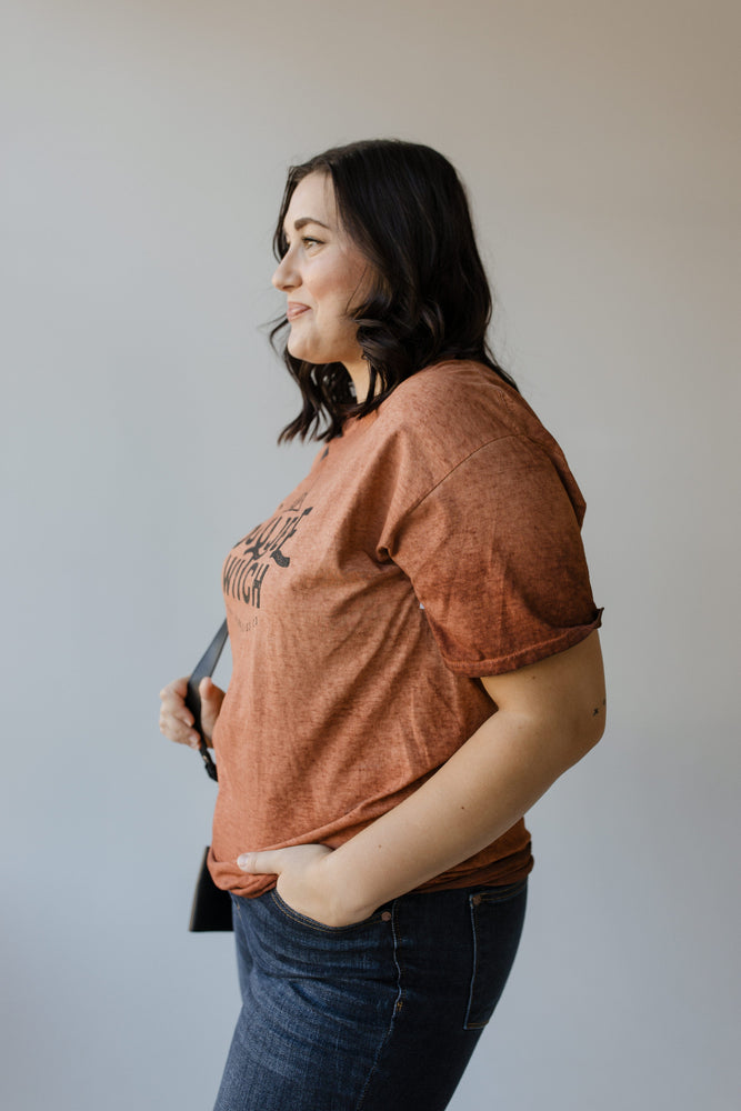 A person with long hair wearing a brown "BOUJEE WITCH" GRAPHIC TEE by Love Marlow and a black strap, seen from the back, stands against a neutral background. The shirt adds an air of mysterious elegance to their look.