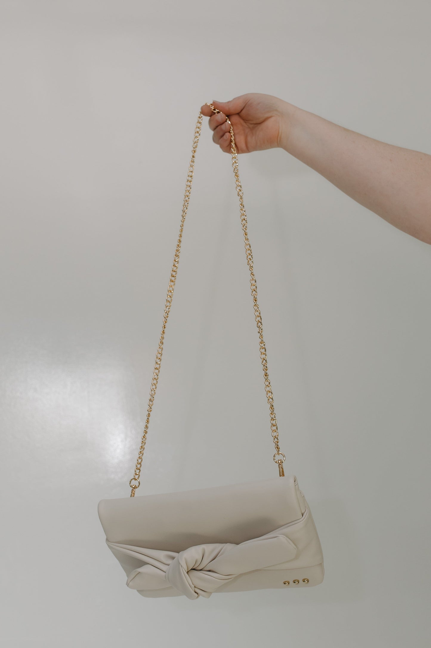 Beige handbag with a gold chain held by a hand against a plain background