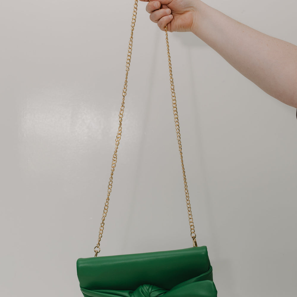 Green handbag with a gold chain strap held by a hand against a plain background