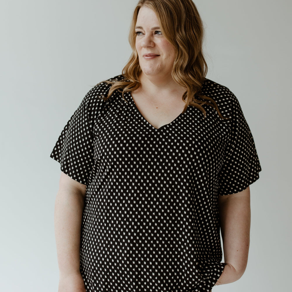 The individual with wavy hair is wearing the Yesta BREEZY ABSTRACT DOT BLOUSE in black and white, paired with beige pants, standing gracefully against a plain background.