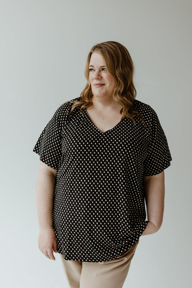 The individual with wavy hair is wearing the Yesta BREEZY ABSTRACT DOT BLOUSE in black and white, paired with beige pants, standing gracefully against a plain background.