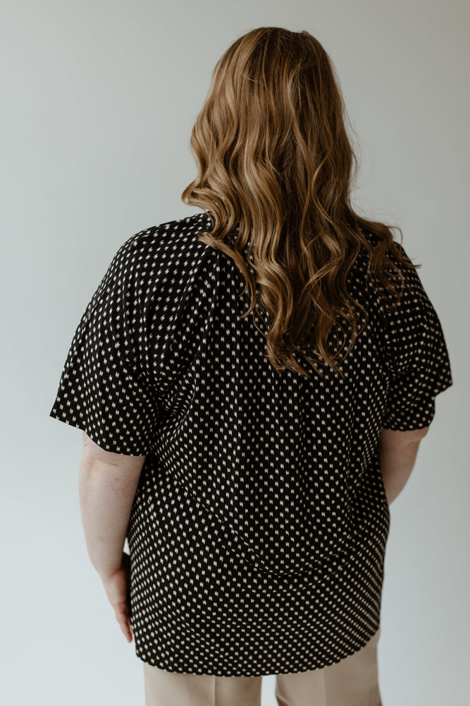 A person with long wavy hair is wearing the Yesta Breezy Abstract Dot Blouse in black and white, perfectly paired with wrinkle-free beige pants, while standing with their back to the camera.