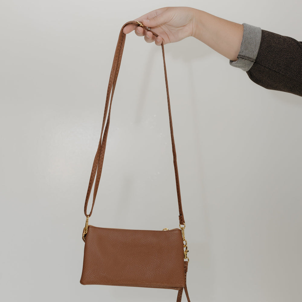Brown leather crossbody bag held by a hand against a plain background
