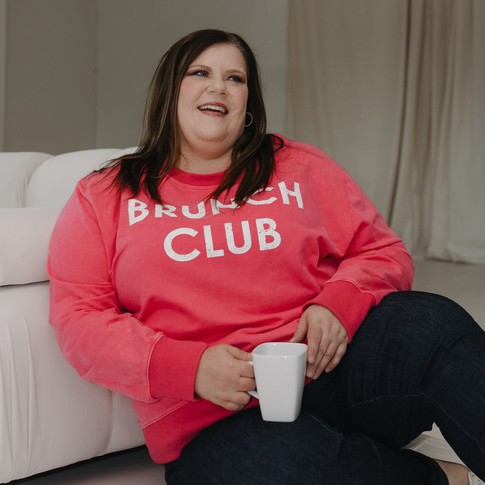 Woman wearing a 'Brunch Club' sweatshirt sitting on a couch holding a mug.