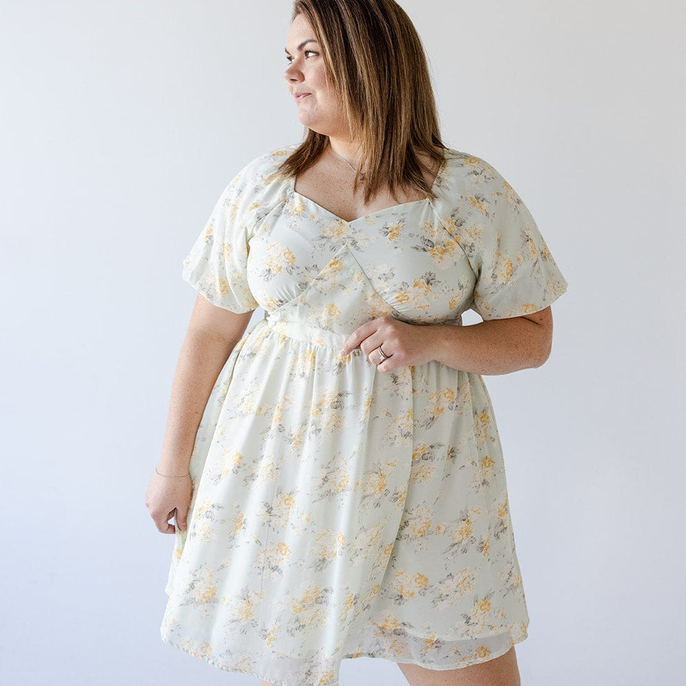 A woman with brown hair is standing against a plain background, wearing the Love Marlow BUBBLE SLEEVE A-LINE DRESS IN SUNRISE YELLOW. She is looking to her left with one hand resting by her side and the other holding the dress.