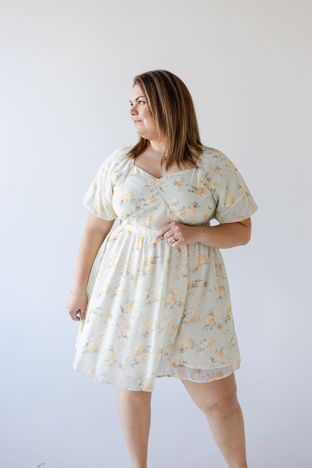 A woman with brown hair is standing against a plain background, wearing the Love Marlow BUBBLE SLEEVE A-LINE DRESS IN SUNRISE YELLOW. She is looking to her left with one hand resting by her side and the other holding the dress.