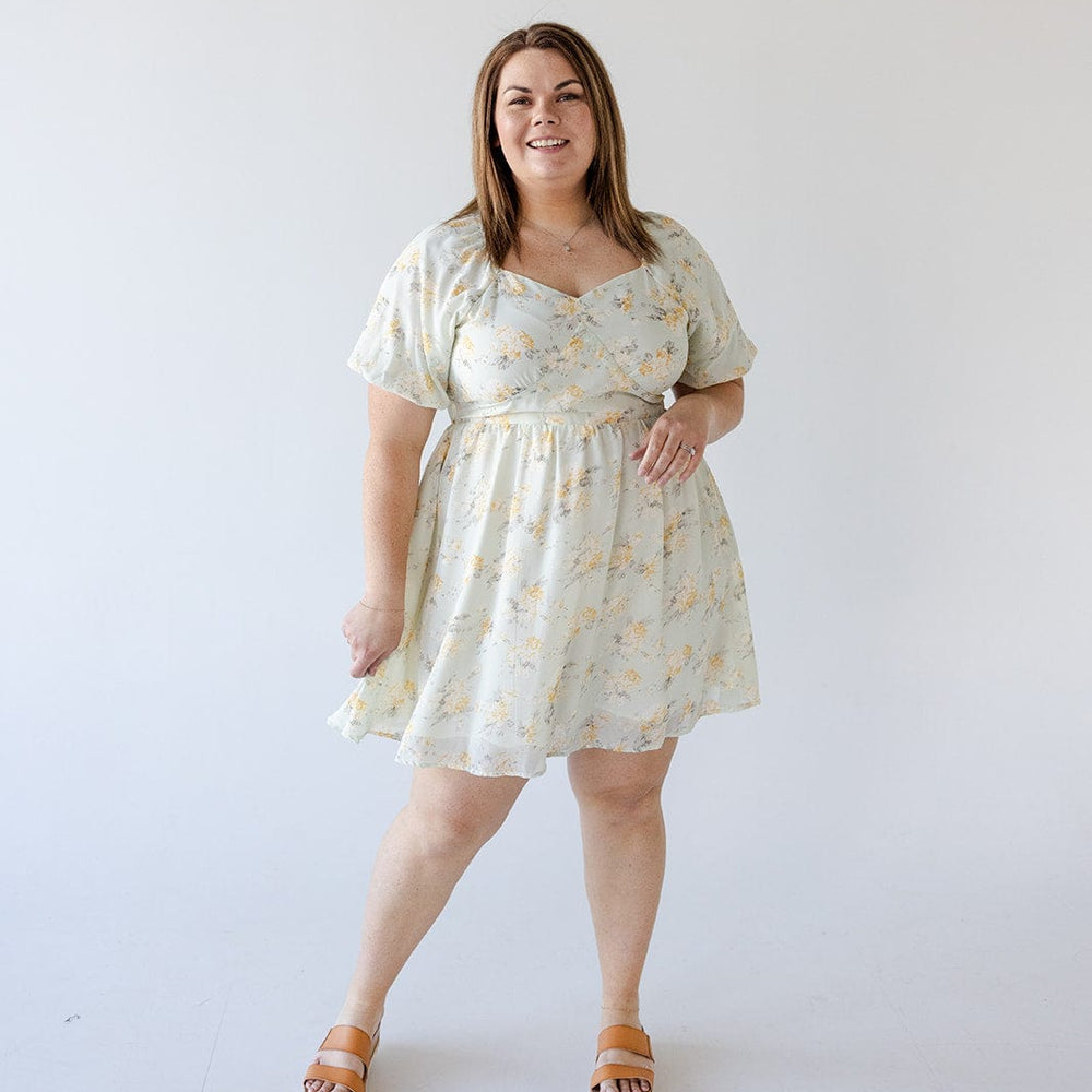 A woman stands against a plain background wearing a knee-length BUBBLE SLEEVE A-LINE DRESS IN SUNRISE YELLOW by Love Marlow with pockets and sandals. She has a neutral expression and one hand by her side.