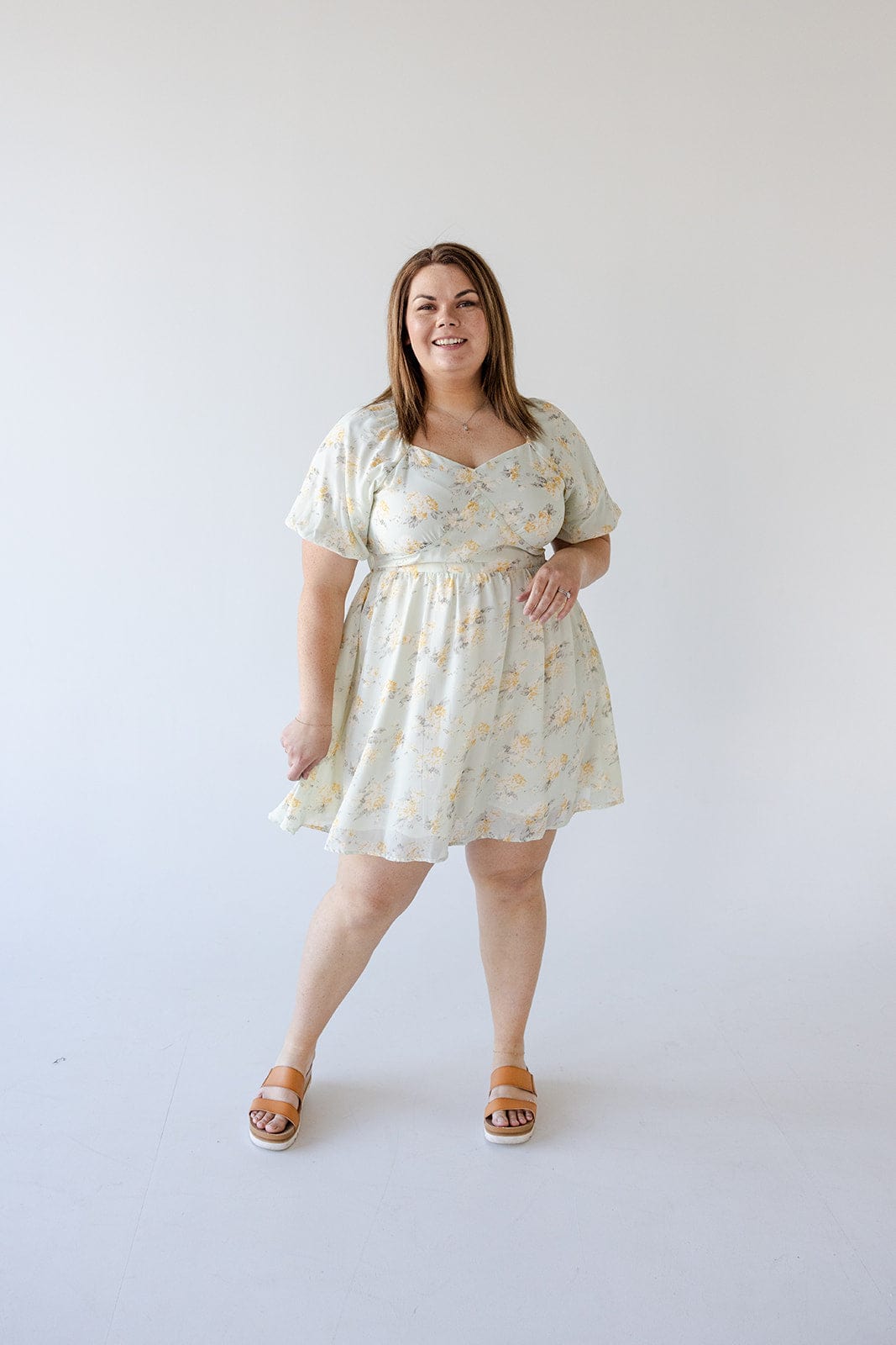 A woman stands against a plain background wearing a knee-length BUBBLE SLEEVE A-LINE DRESS IN SUNRISE YELLOW by Love Marlow with pockets and sandals. She has a neutral expression and one hand by her side.