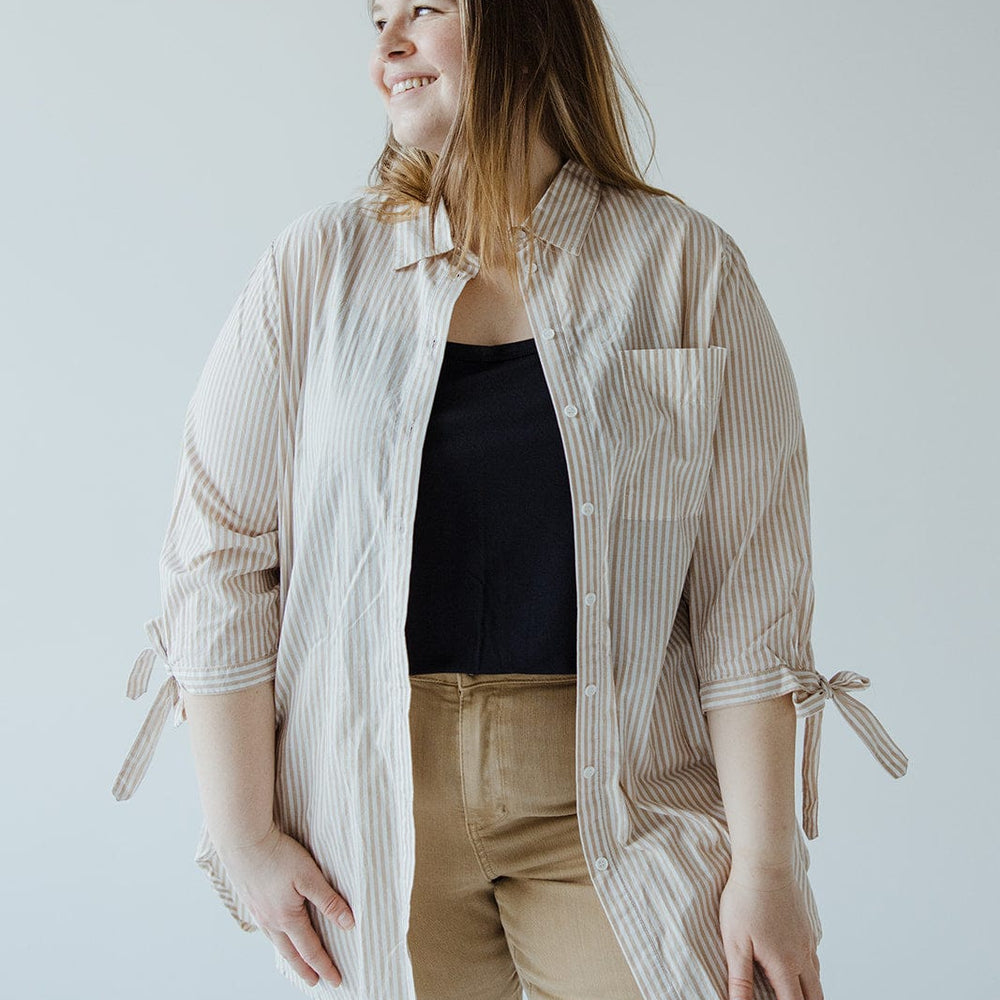 A person wearing a Love Marlow BUTTON-UP COLLARED TUNIC BLOUSE IN SAND that features summer stripes over a black top and beige pants, smiling and looking to the side.