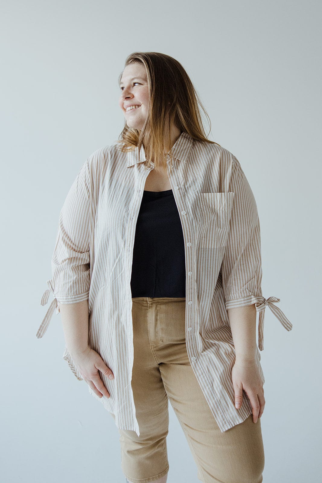 A person wearing a Love Marlow BUTTON-UP COLLARED TUNIC BLOUSE IN SAND that features summer stripes over a black top and beige pants, smiling and looking to the side.