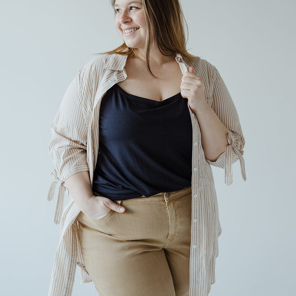 A woman stands smiling, wearing a striped shirt over a black top and beige pants, with one hand in her pocket. The BUTTON-UP COLLARED TUNIC BLOUSE IN SAND by Love Marlow effortlessly complements her outfit.