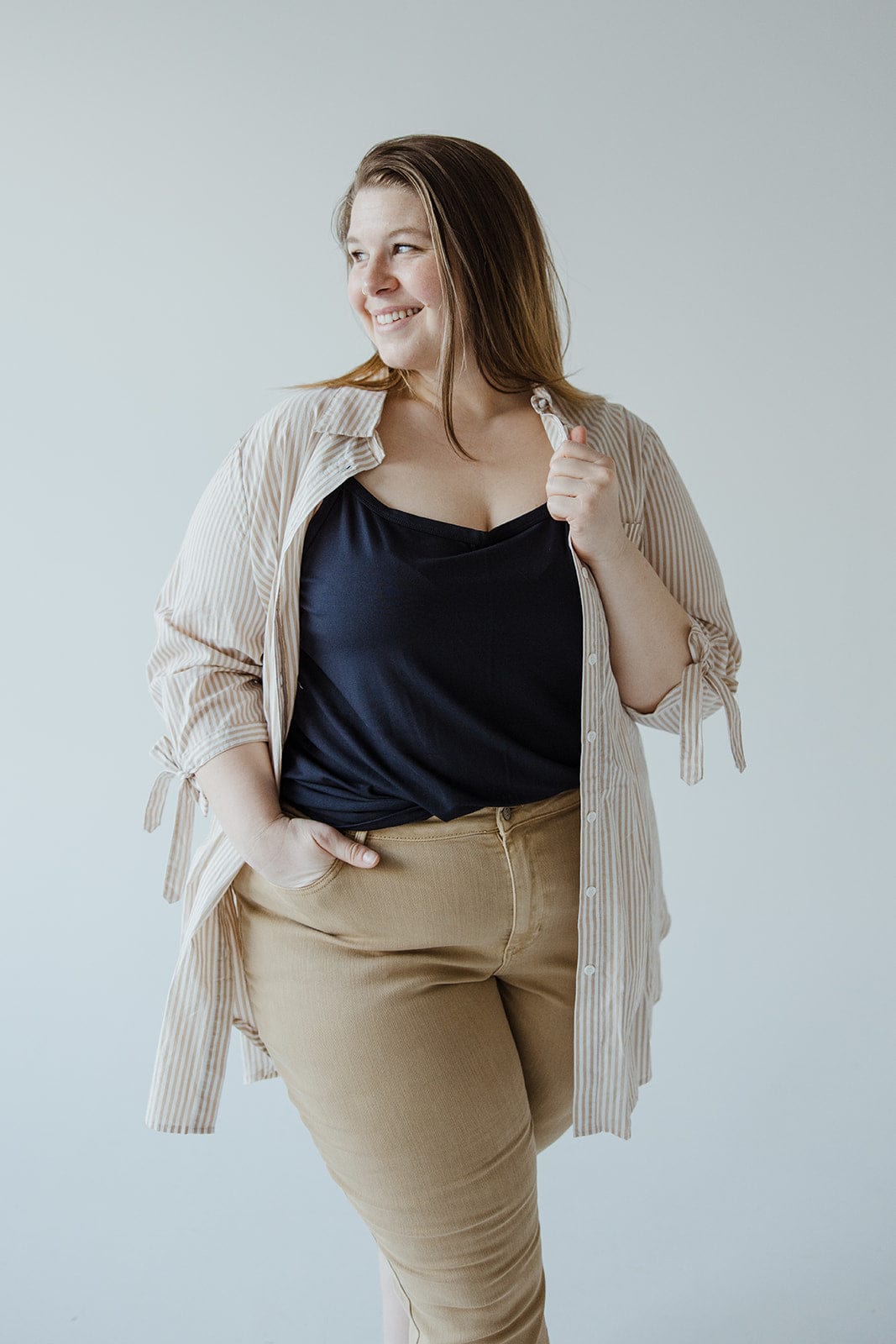 A woman stands smiling, wearing a striped shirt over a black top and beige pants, with one hand in her pocket. The BUTTON-UP COLLARED TUNIC BLOUSE IN SAND by Love Marlow effortlessly complements her outfit.