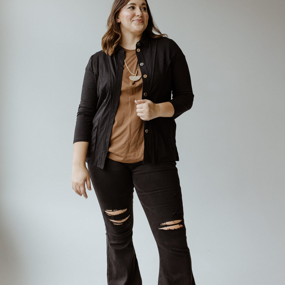 A person wearing the Button-Up Blouse with Raw Seam Details in Black by neon buddha over a flattering fit brown top and ripped black jeans stands smiling against a plain background.