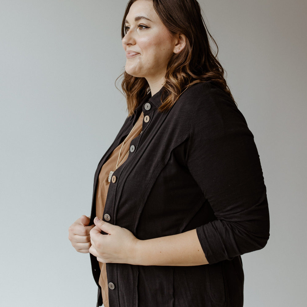 A woman stands sideways, looking ahead. Her BUTTON-UP BLOUSE WITH RAW SEAM DETAILS IN BLACK from neon buddha elegantly layers over a brown blouse, offering a flattering fit against her wavy brown hair. The background remains plain gray.