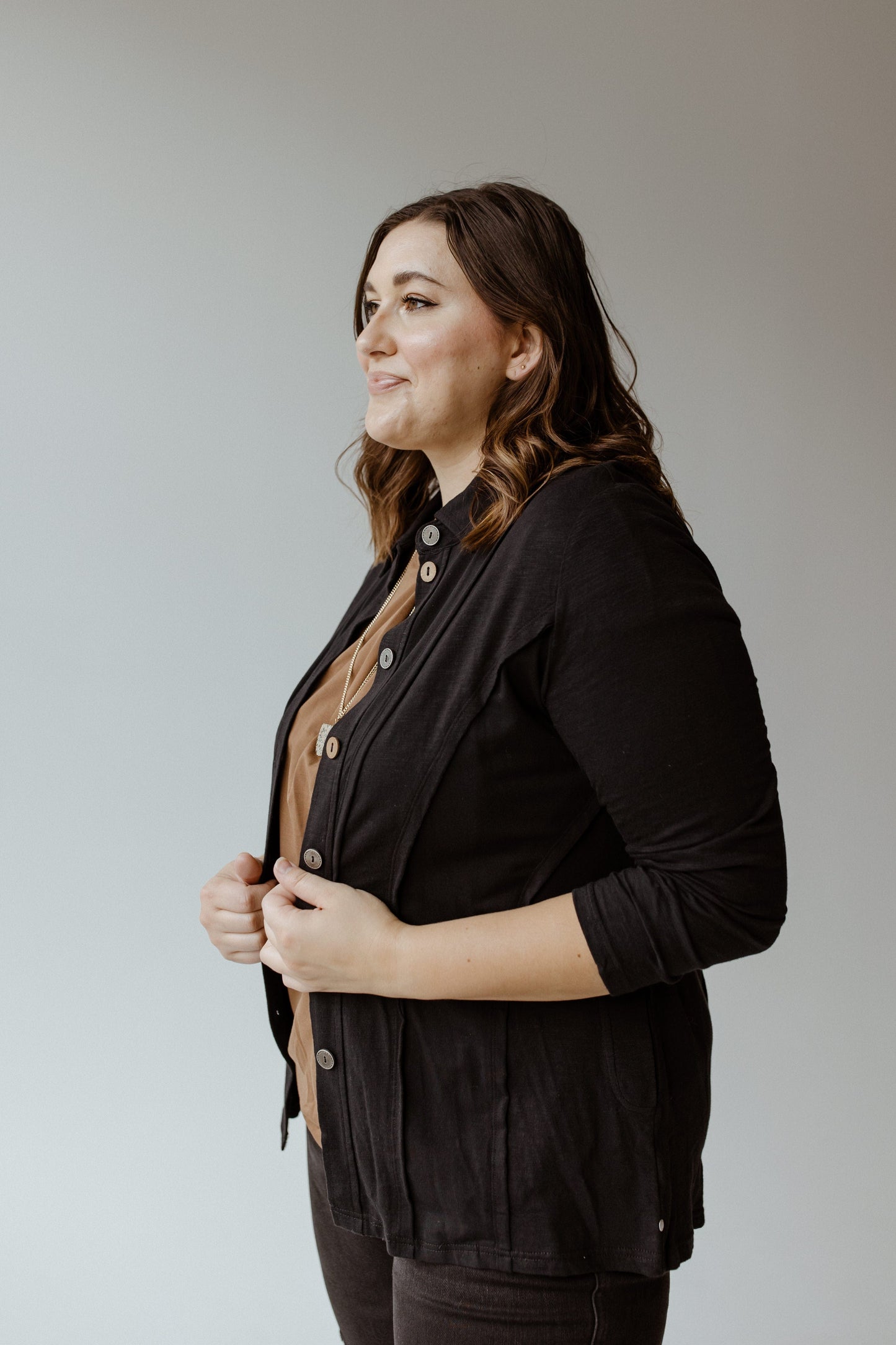 A woman stands sideways, looking ahead. Her BUTTON-UP BLOUSE WITH RAW SEAM DETAILS IN BLACK from neon buddha elegantly layers over a brown blouse, offering a flattering fit against her wavy brown hair. The background remains plain gray.