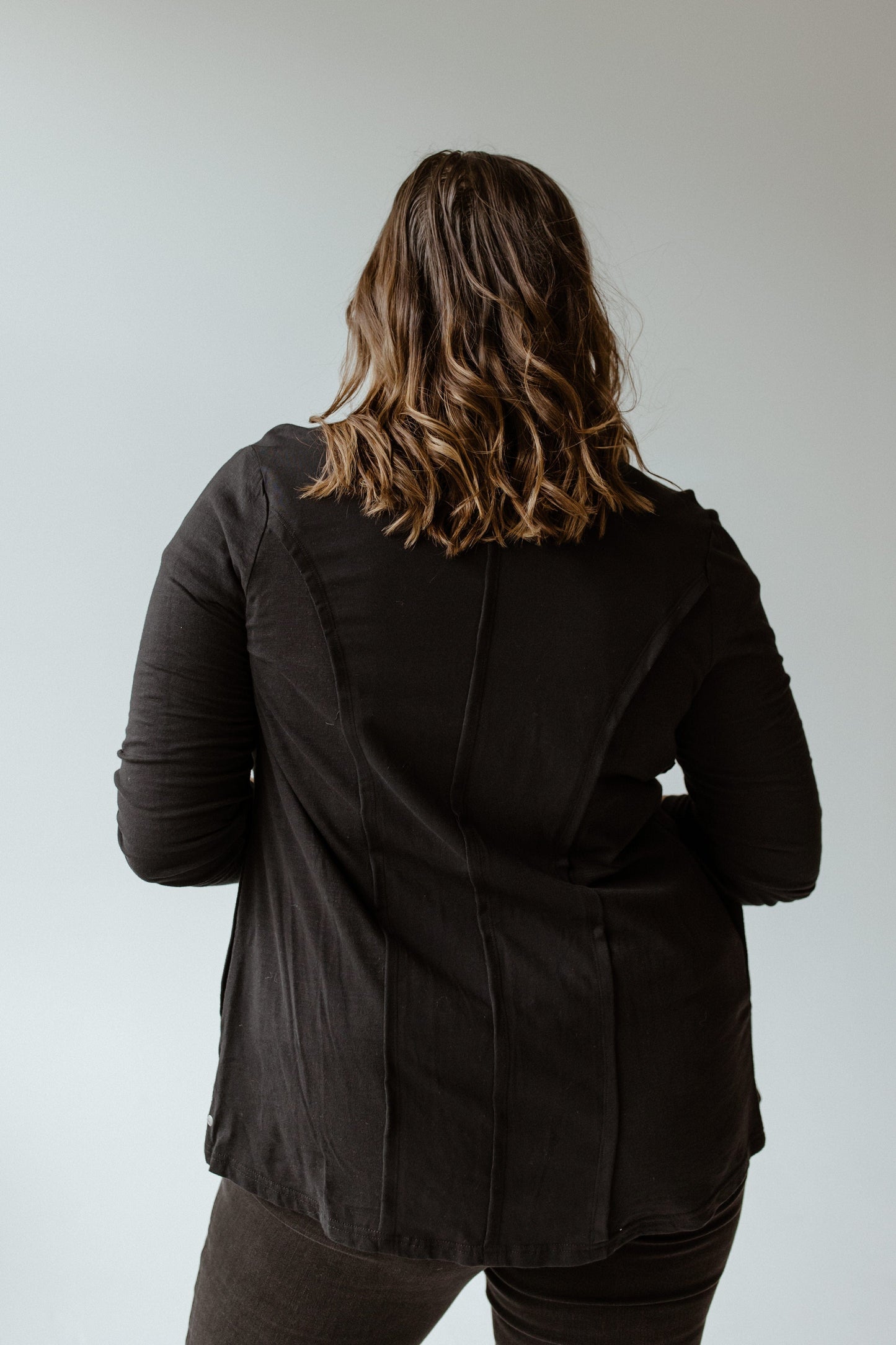 A person with wavy brown hair stands with their back to the camera, highlighting a flattering fit in neon buddha's black Button-Up Blouse with Raw Seam Details and matching pants.