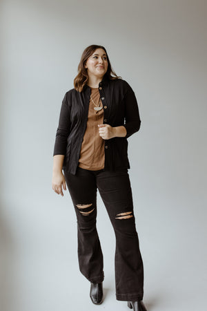 A person wearing the Button-Up Blouse with Raw Seam Details in Black by neon buddha over a flattering fit brown top and ripped black jeans stands smiling against a plain background.