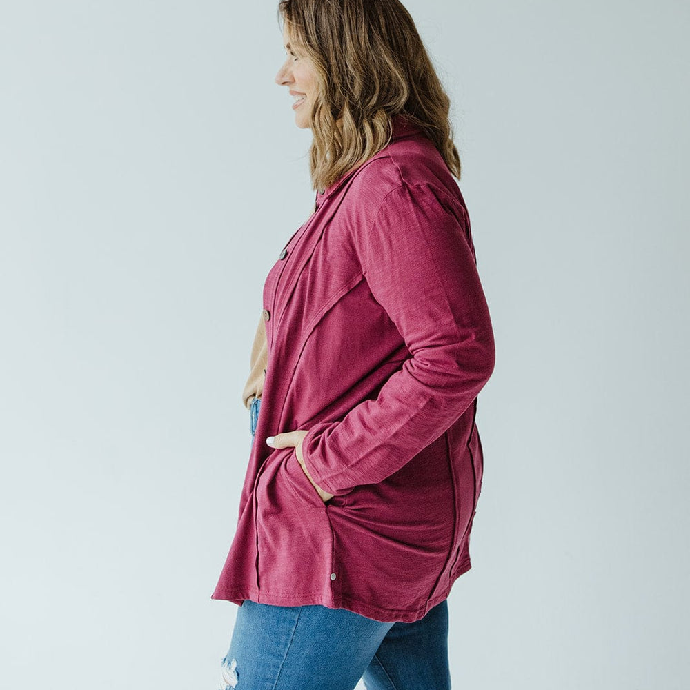 A woman wearing a cranberry button-up blouse with raw seam details from neon buddha and blue jeans stands in a light gray room, facing left with her hands in her pockets.