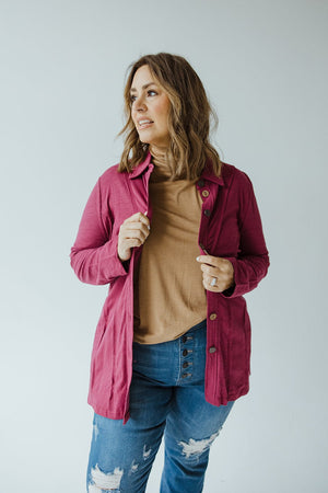 A person with shoulder-length hair wears a pink cardigan over a cranberry button-up blouse with raw seam details from neon buddha, paired with blue jeans. They are facing slightly to the side, adjusting the cardigan with one hand.