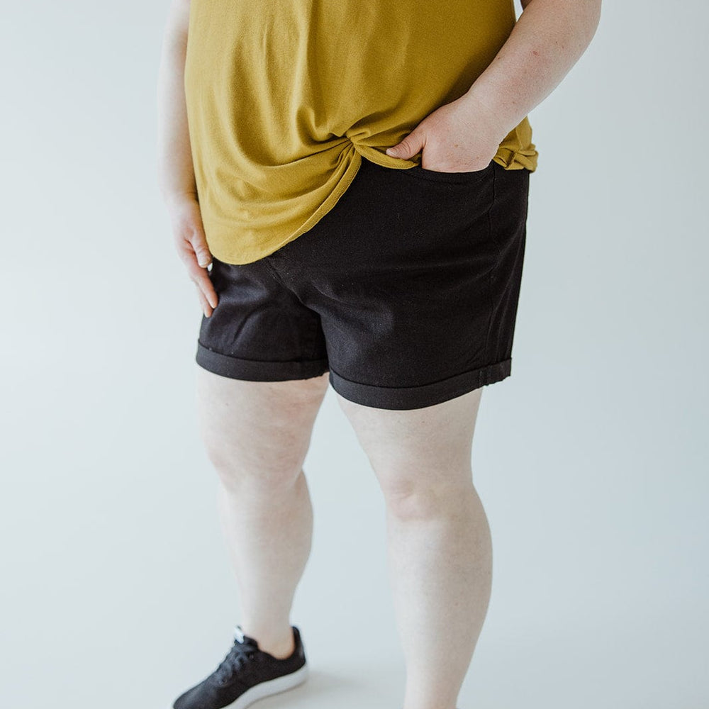 A person wearing a yellow shirt, BUTTON FLY CUFFED SHORTS IN BLACK by Love Marlow, and black sneakers stands indoors. The person's hand is partially tucked into a pocket.