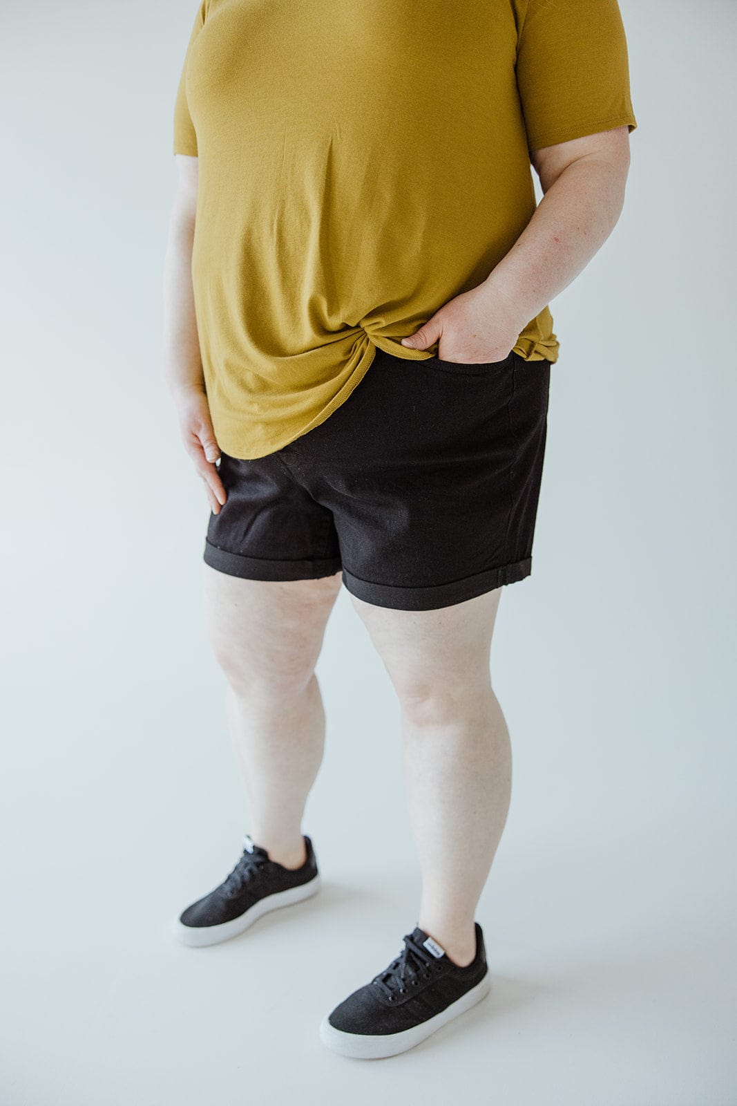 A person wearing a yellow shirt, BUTTON FLY CUFFED SHORTS IN BLACK by Love Marlow, and black sneakers stands indoors. The person's hand is partially tucked into a pocket.