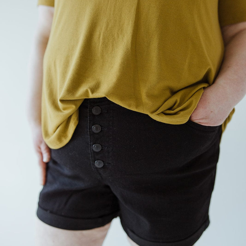 A person wears a mustard yellow top and Love Marlow BUTTON FLY CUFFED SHORTS IN BLACK, standing with one hand in their pocket against a neutral background.