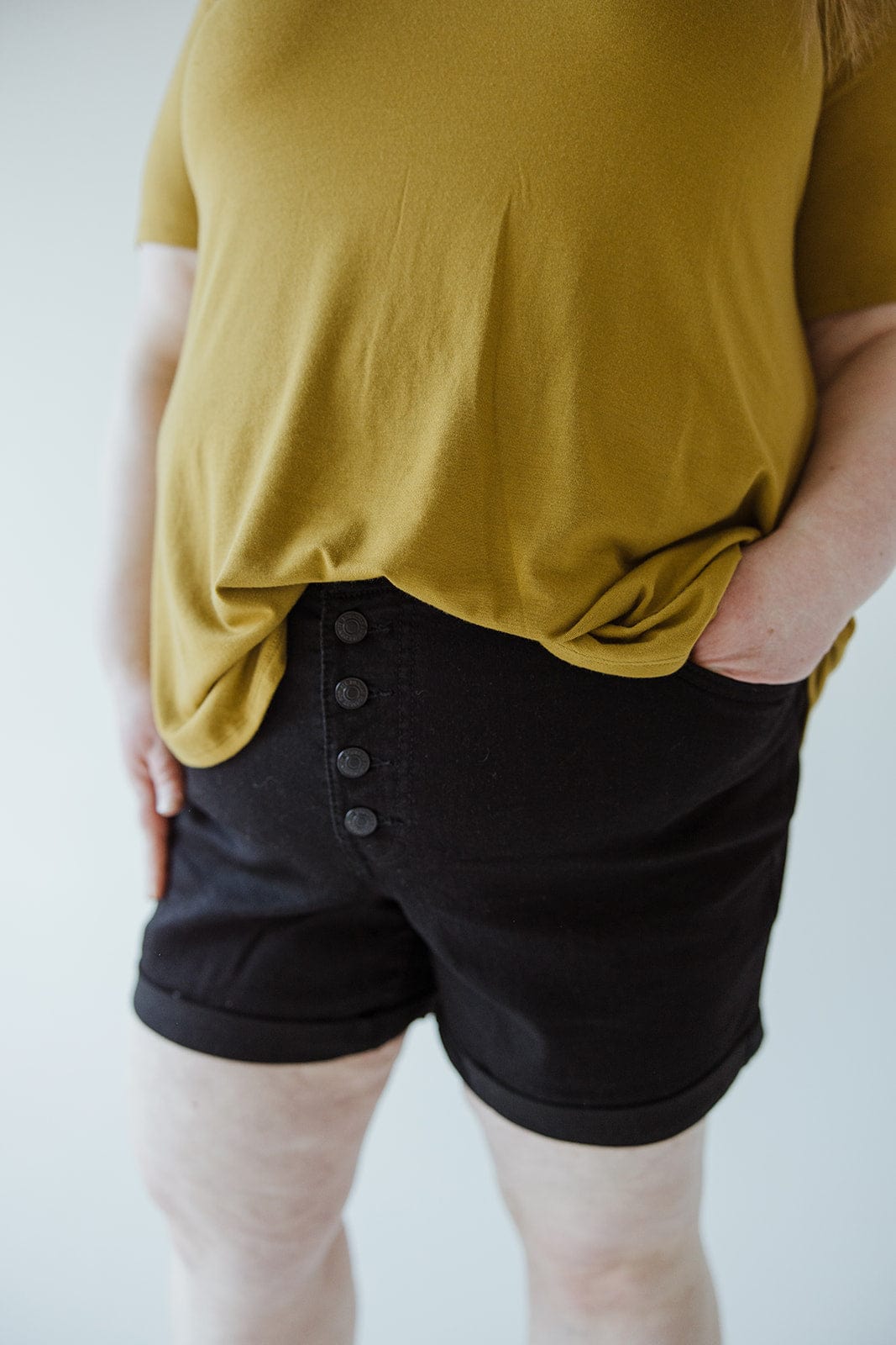 A person wears a mustard yellow top and Love Marlow BUTTON FLY CUFFED SHORTS IN BLACK, standing with one hand in their pocket against a neutral background.