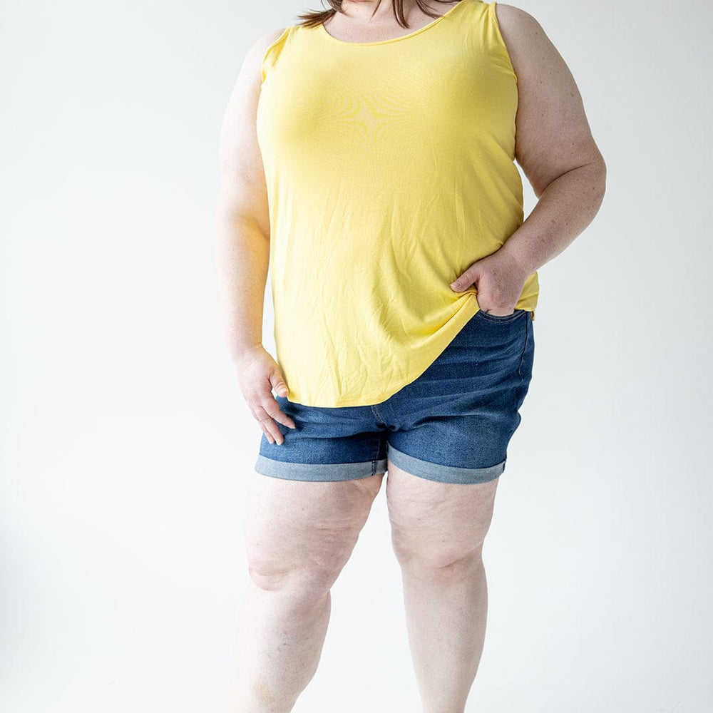 A woman stands against a neutral background wearing a yellow sleeveless top, BUTTON FLY CUFFED SHORTS IN DARK INDIGO by Love Marlow, and black sandals. She has her left hand in her pocket and looks to the side.