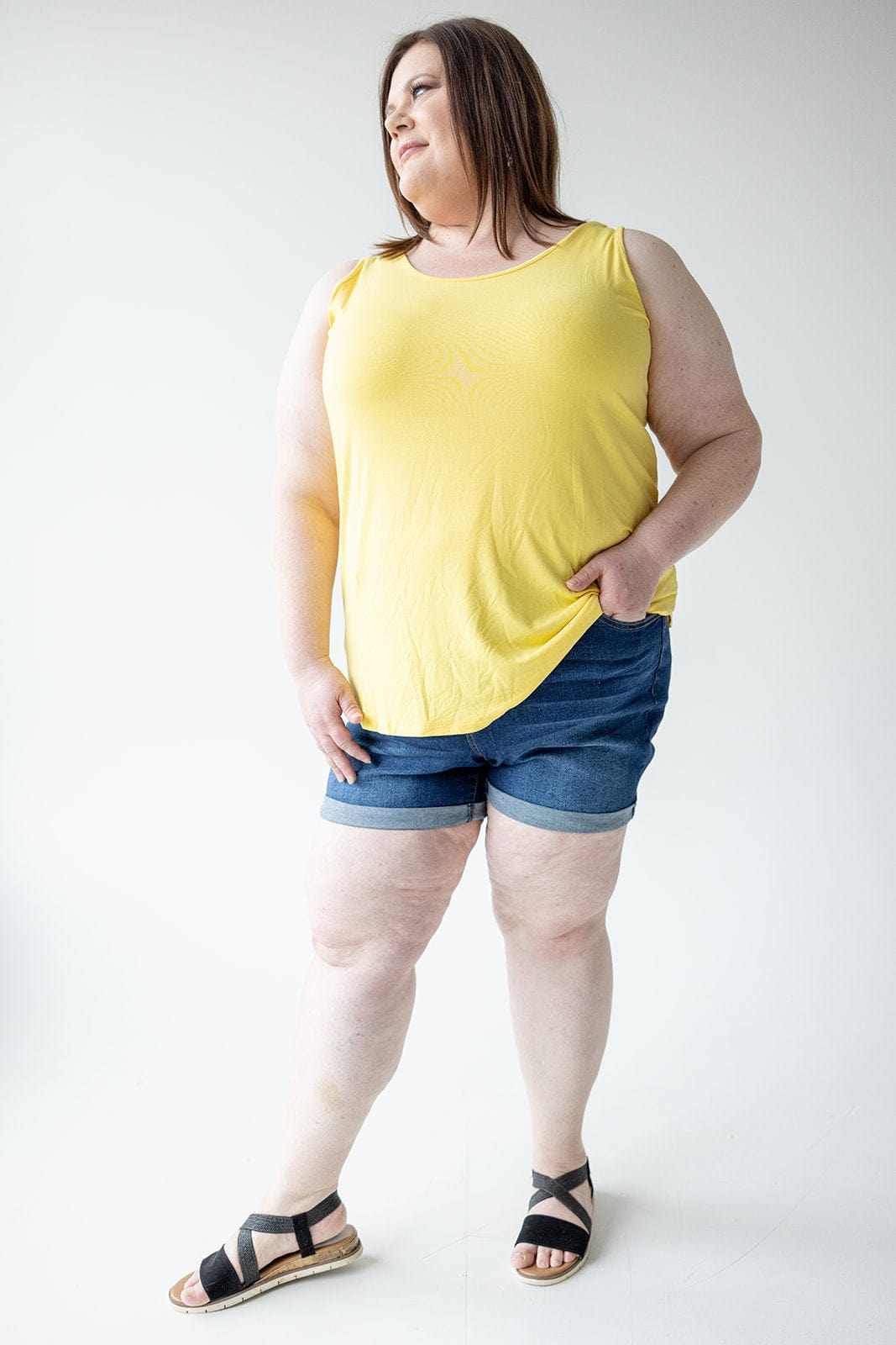 A woman stands against a neutral background wearing a yellow sleeveless top, BUTTON FLY CUFFED SHORTS IN DARK INDIGO by Love Marlow, and black sandals. She has her left hand in her pocket and looks to the side.