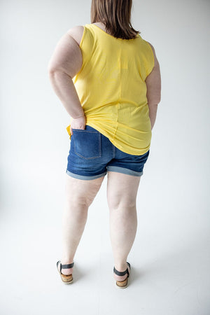 A person with a yellow sleeveless top and BUTTON FLY CUFFED SHORTS IN DARK INDIGO from Love Marlow stands facing a neutral background, with one hand placed in a back pocket, visible from the back.