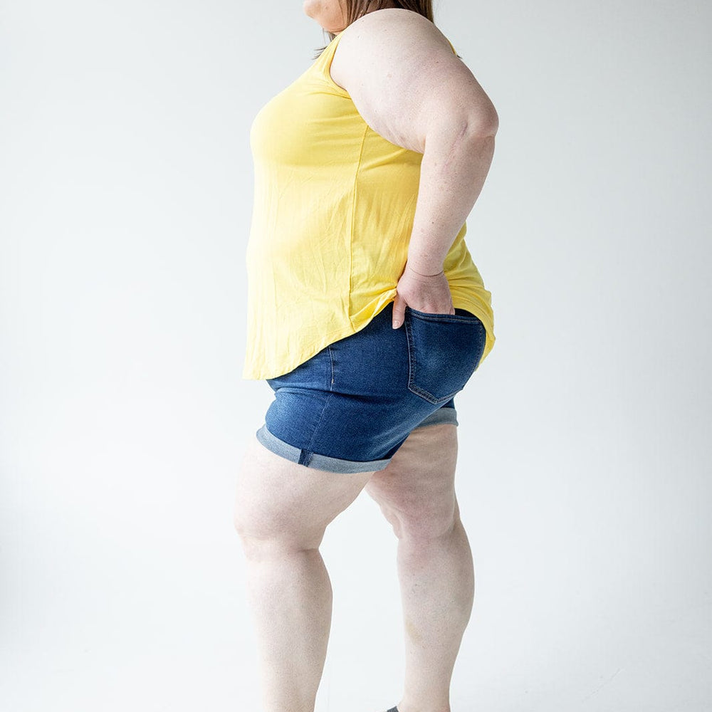 A person stands in profile, wearing a yellow sleeveless top, Love Marlow BUTTON FLY CUFFED SHORTS IN DARK INDIGO, and black sandals against a plain white background.