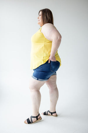 A person stands in profile, wearing a yellow sleeveless top, Love Marlow BUTTON FLY CUFFED SHORTS IN DARK INDIGO, and black sandals against a plain white background.