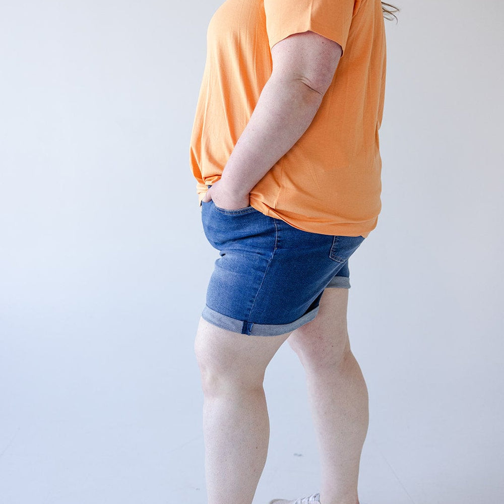 Person standing sideways against a plain background, wearing an orange T-shirt, Love Marlow BUTTON FLY CUFFED SHORTS IN MEDIUM WASH, and light-colored sneakers with hands in pockets.