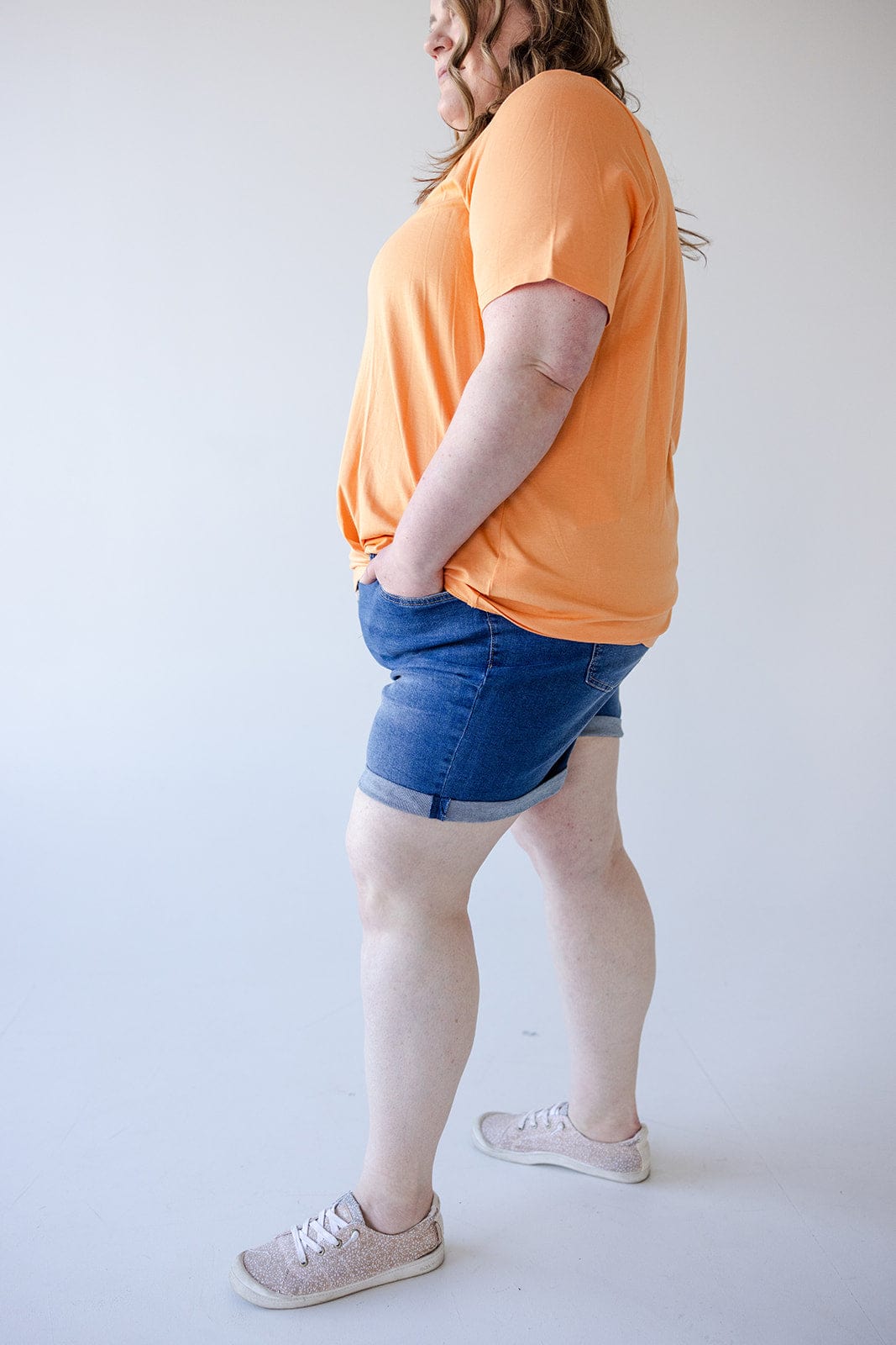 Person standing sideways against a plain background, wearing an orange T-shirt, Love Marlow BUTTON FLY CUFFED SHORTS IN MEDIUM WASH, and light-colored sneakers with hands in pockets.