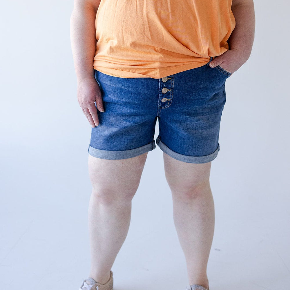 Person wearing an orange shirt, BUTTON FLY CUFFED SHORTS IN MEDIUM WASH by Love Marlow with 5 pocket construction, and beige sneakers, standing with one hand in pocket.